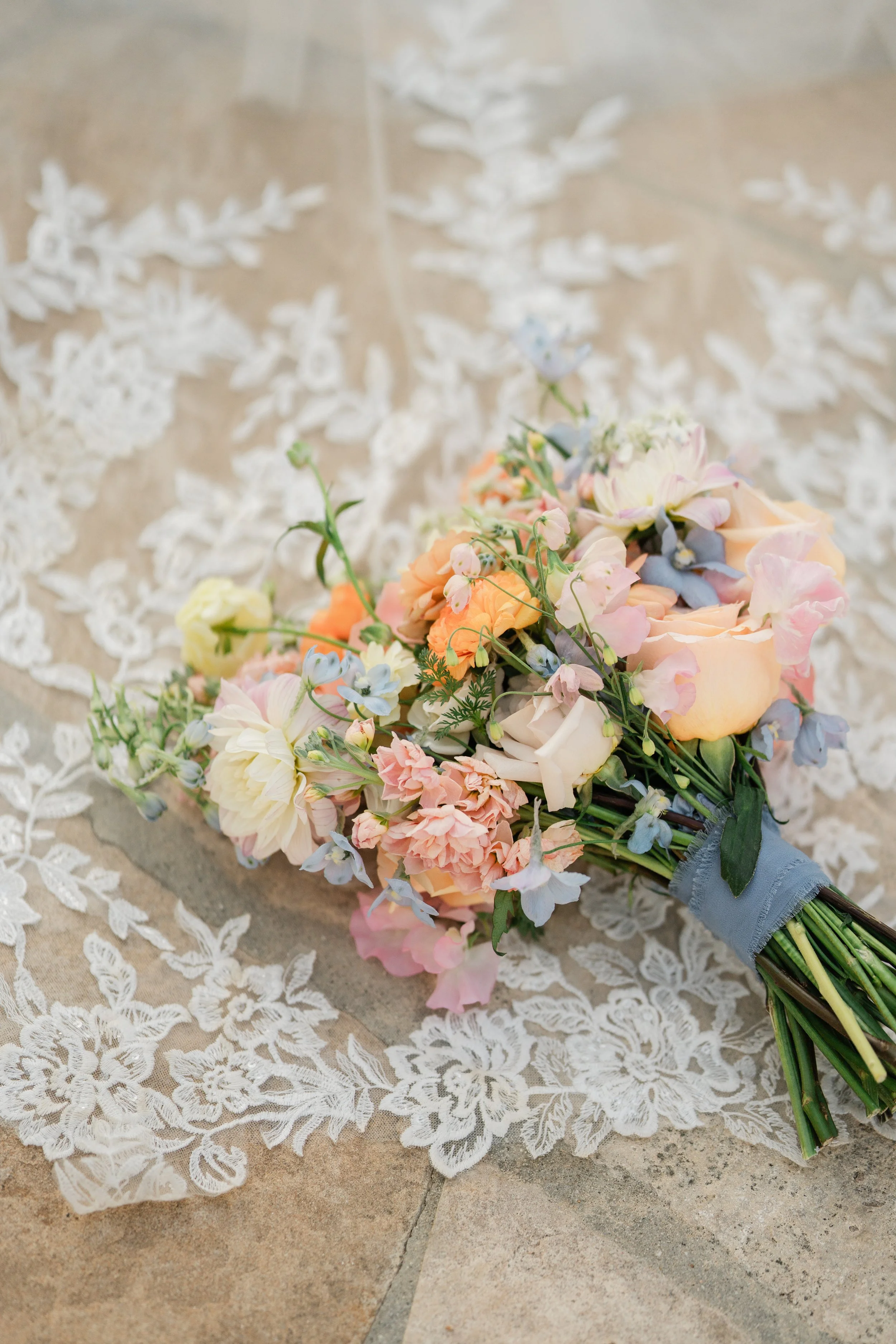 A colorful bouquet of pastel flowers, including roses, hydrangeas, and other blossoms, lies on lace fabric on a tiled floor.