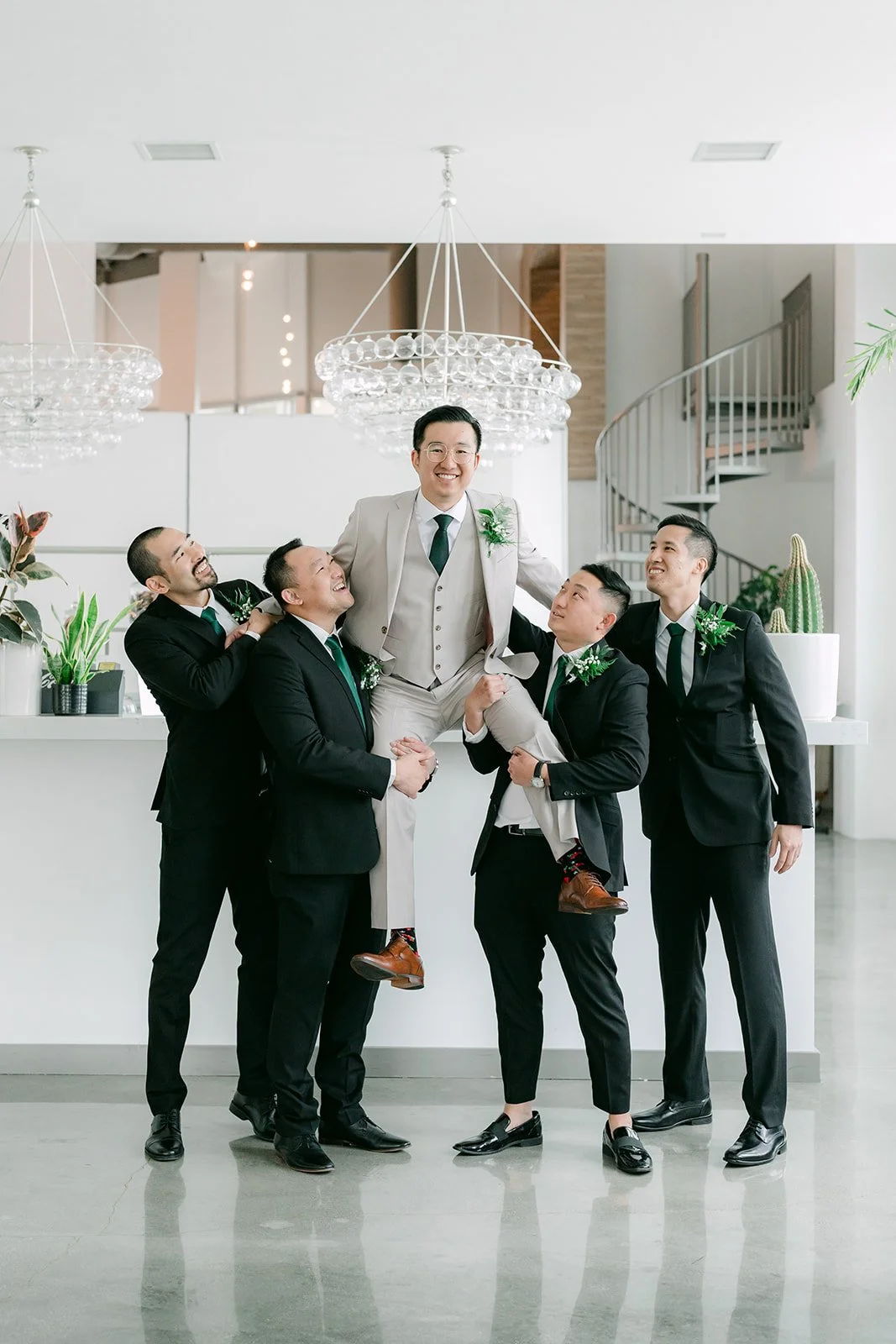 Groomsmen in black suits lifting Ricky on a white staircase