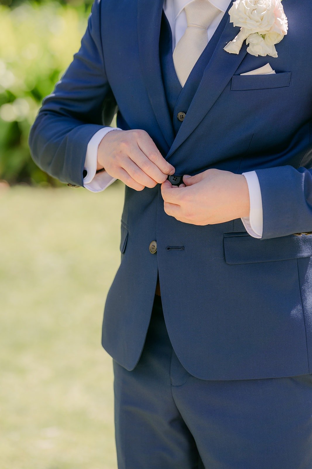 Alex buttoning his navy jacket, with a white rose boutonniere on his lapel