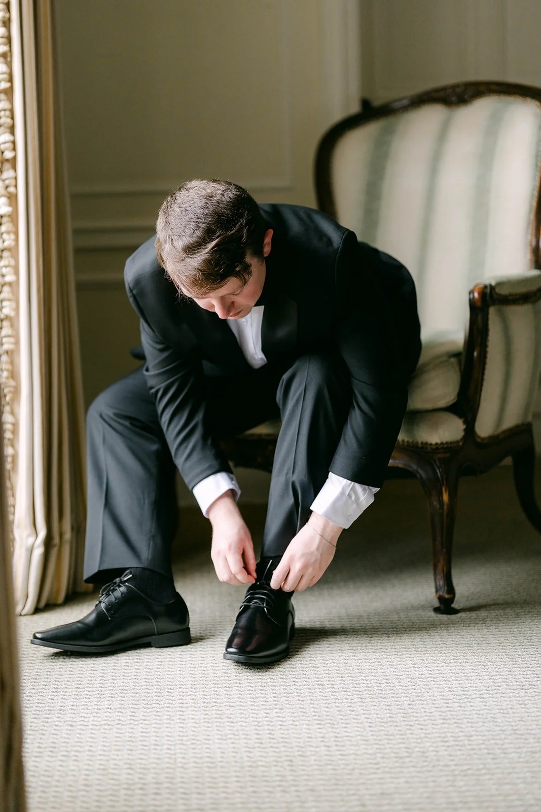 Matthew tying his dress shoes in an upholstered armchair