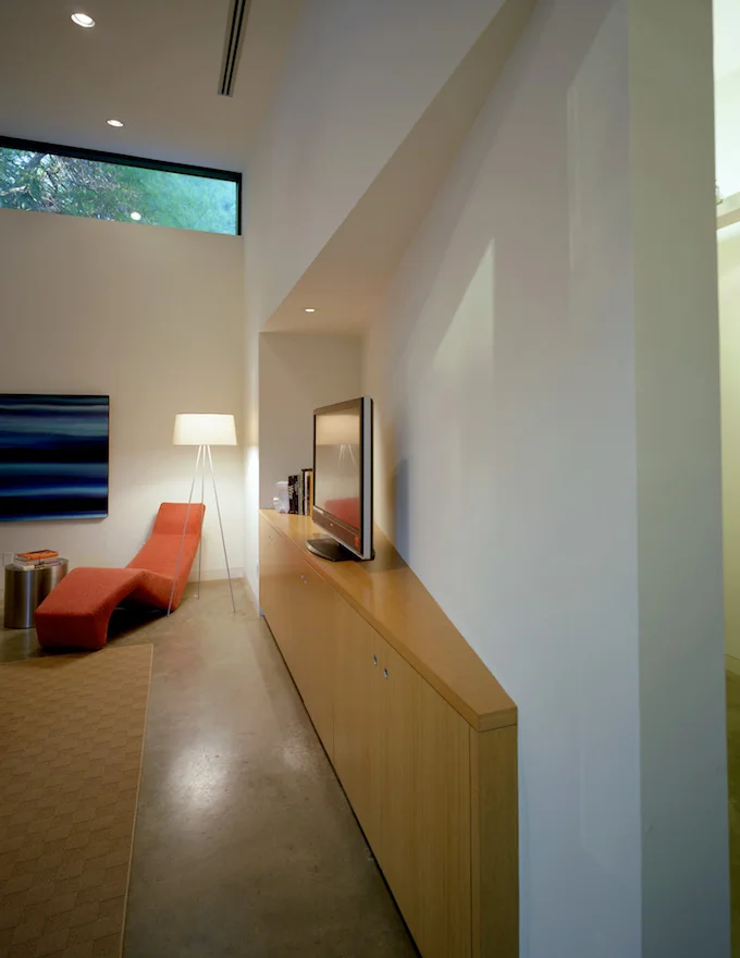 The skewed walls and varied openings provide a drama reminiscent of narrow canyons or ancient ruins. The living areas open to buffalo grass, live oaks, and terraces of indigenous plants. A rugged coursed stone and paint-grip galvanized metal roof contrast with the sharply carved interior of honed concrete floors, white walls and white oak wood elements.