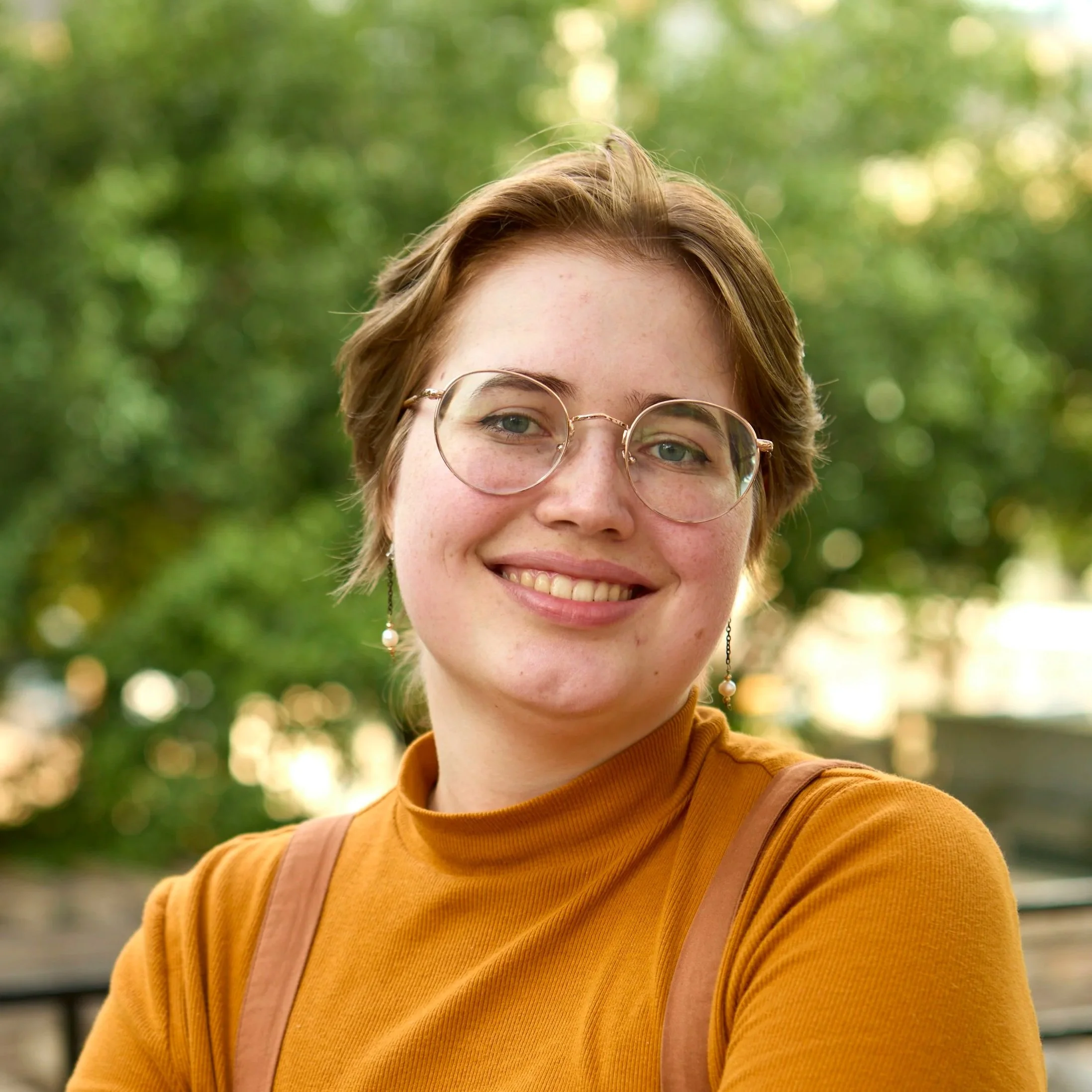 A young person with short brown hair, wearing glasses, earrings, and an orange turtleneck, smiling outdoors with green trees in the background.