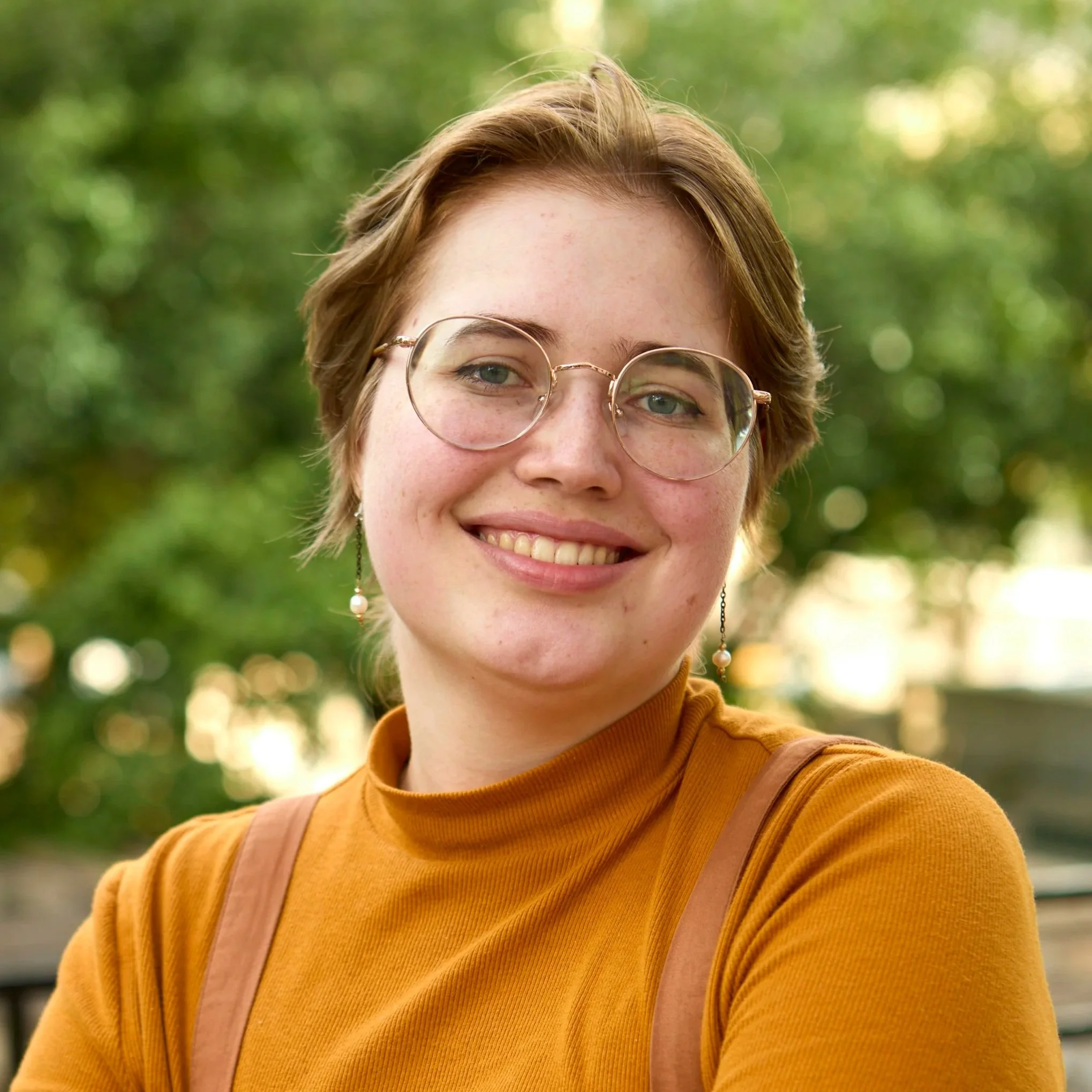 A young person with short brown hair, glasses, and earrings, smiling outdoors in front of green trees, wearing an orange turtleneck.