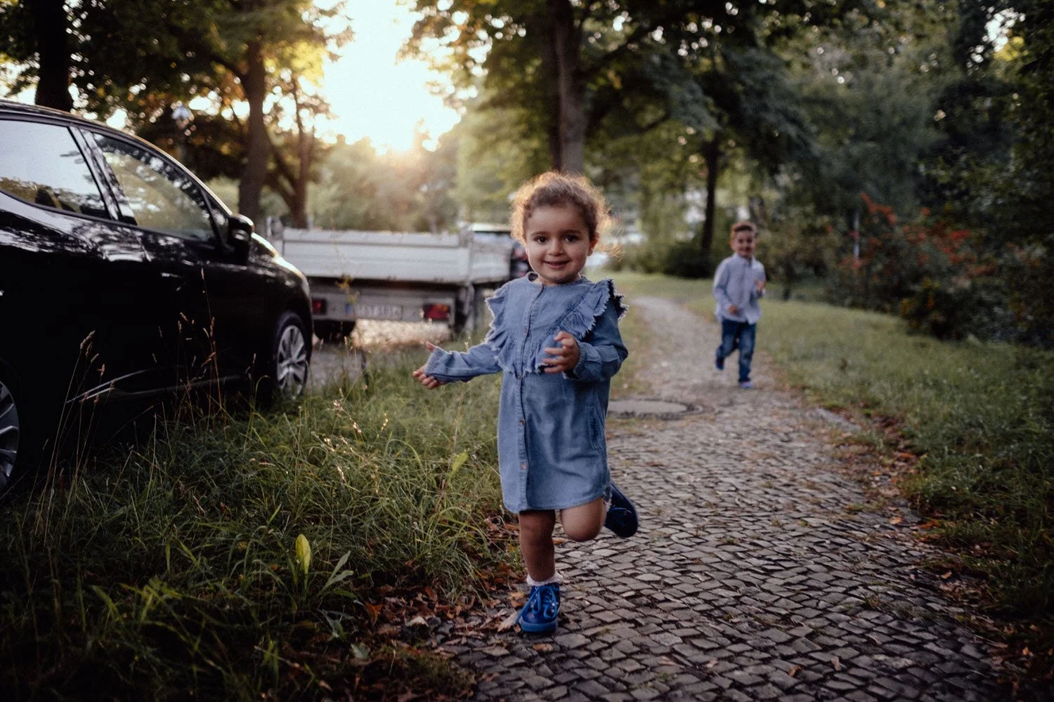 Ein kleines Mädchen und Junge laufen auf einem gepflasterten Weg in Berlin, während die Sonne im Hintergrund untergeht, im Park bei Dämmerung.