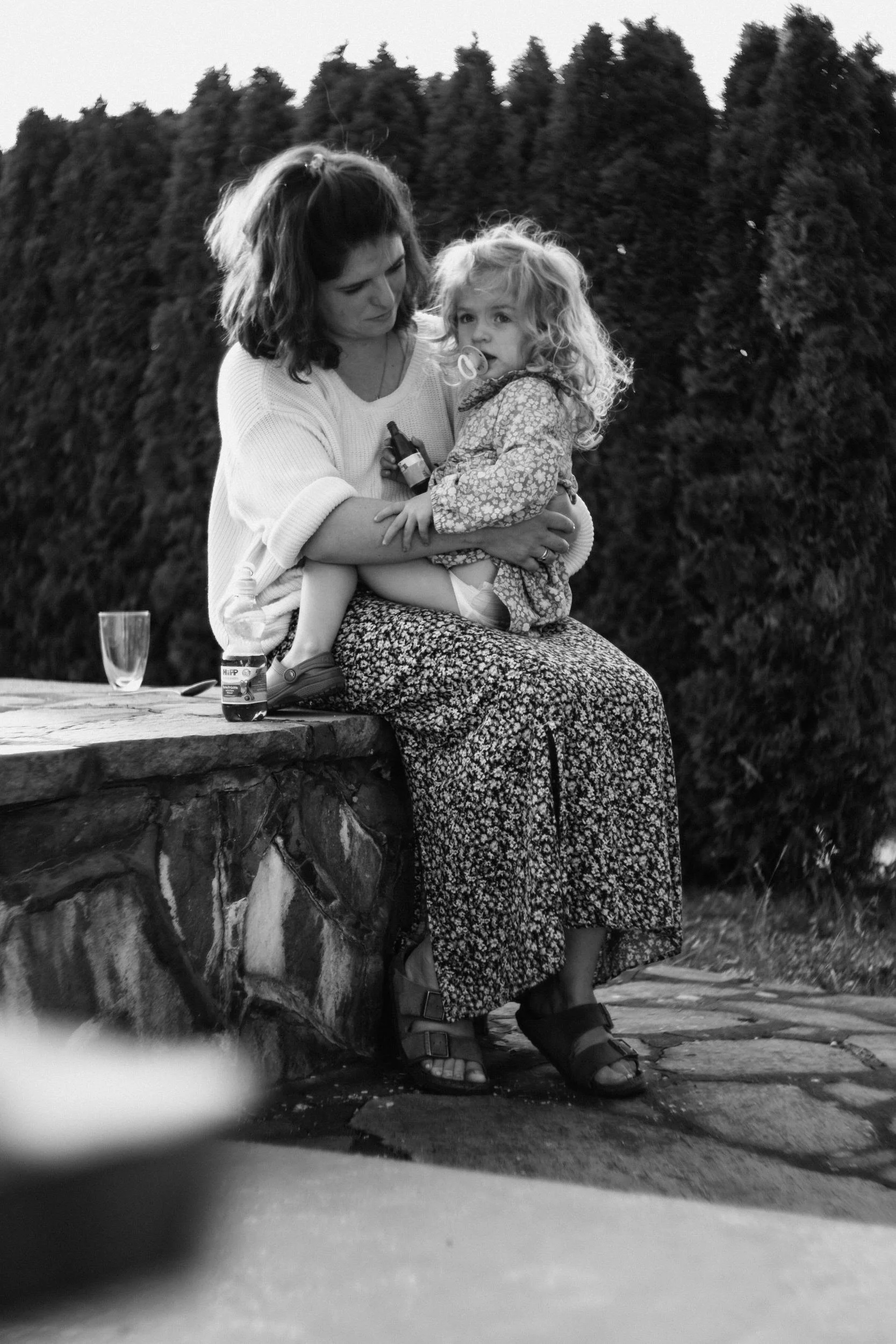 Eine Frau sitzt auf einem Stein im Garten, hält ein kleines Mädchen im Arm. Es ist schwarz-weiß, im Hintergrund ist eine Hecke zu sehen.