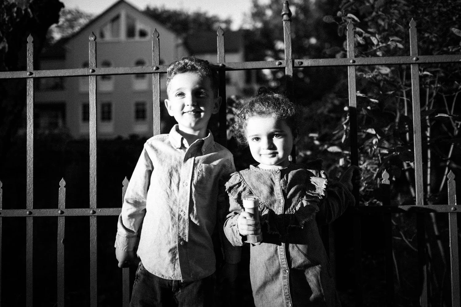 Geschwister, ein Junge und ein Mädchen, stehen vor einem Zaun im Freien bei Sonnenuntergang, mit einem Haus im Hintergrund, das von Licht erleuchtet wird, in schwarz-weiß.