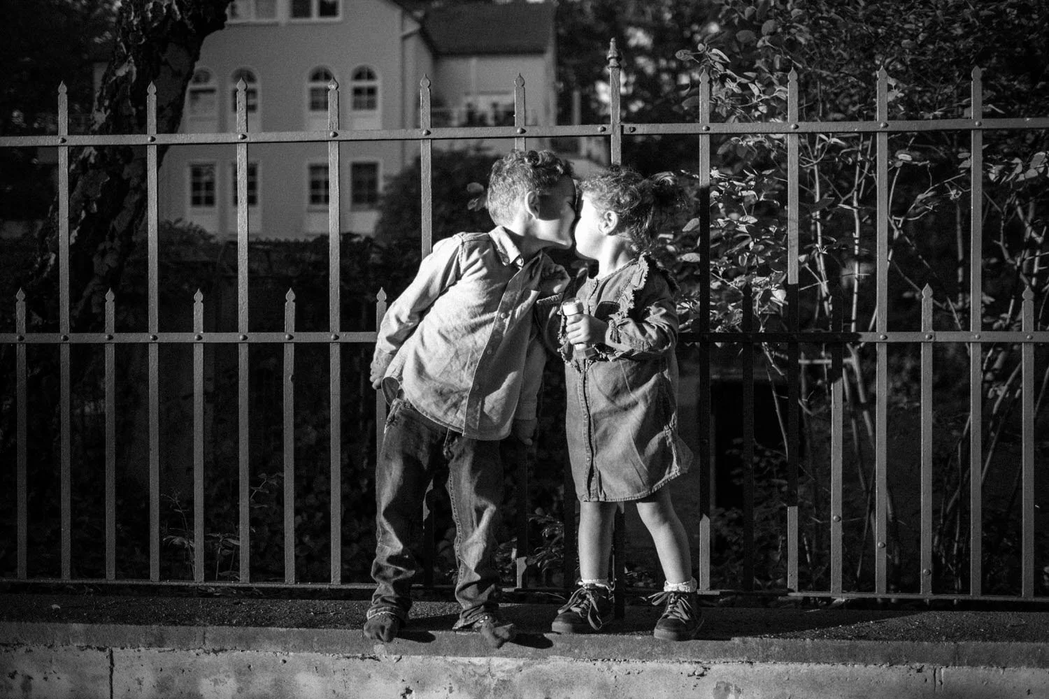 Zwei Kinder küssen sich vor einem Zaun in schwarz-weiß. Eine liebevolle Zuneigung unter Geschwistern.