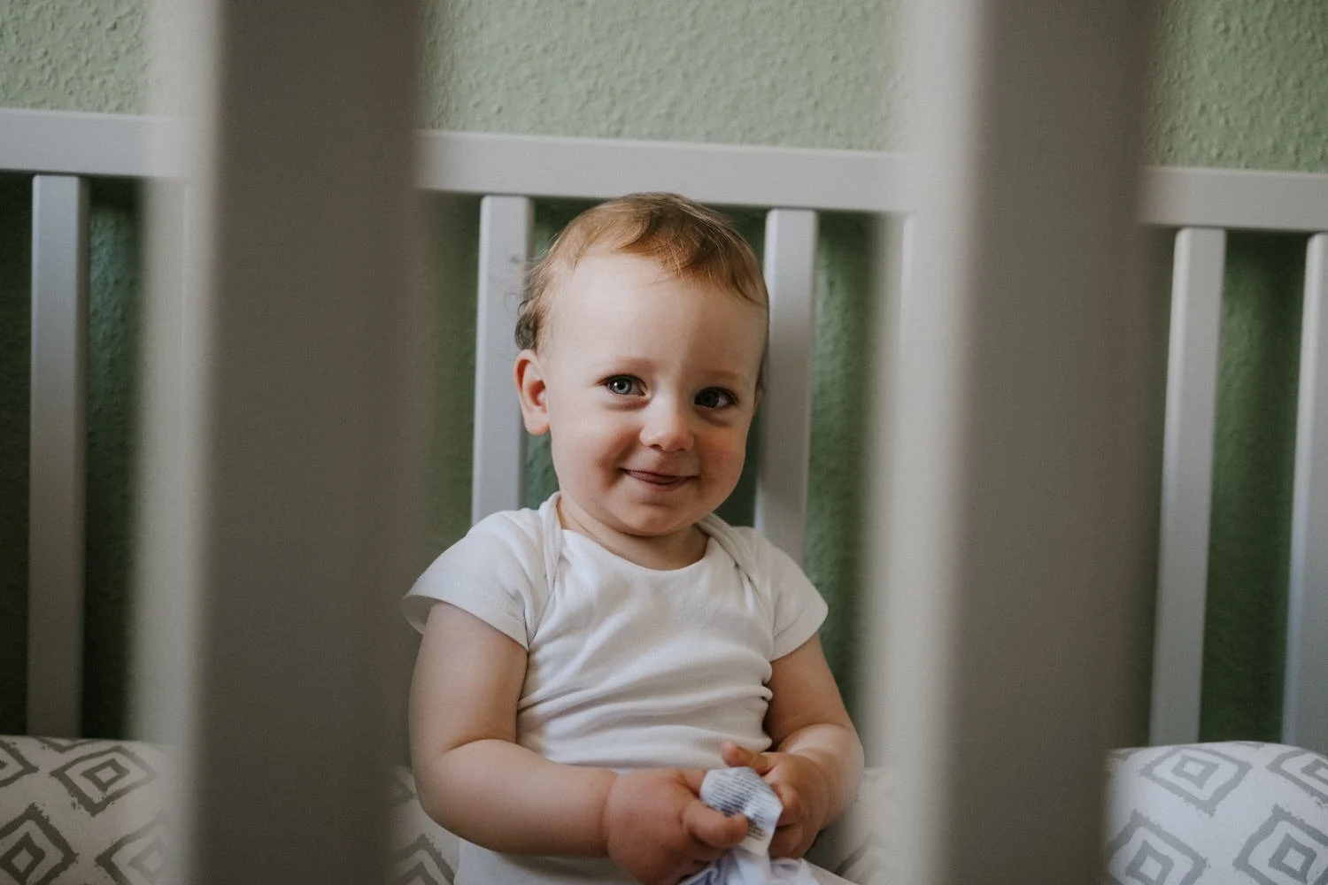 Ein kleiner Junge mit braunen Haaren und blauen Augen sitzt hinter Gitterstäben eines Kinderbetts, trägt ein weißes T-Shirt und lächelt in die Kamera, während er ein kleines Stofftier hält.