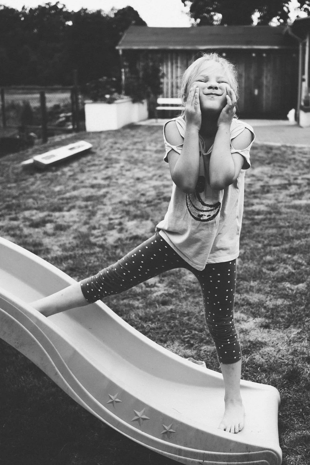 Ein lacheEin lachendes Mädchen steht auf einer kleinen Rutsche im Garten und hält sich in einer lustigen Momentaufnahme die Wangen mit den Händen, während sie eine Pose macht.