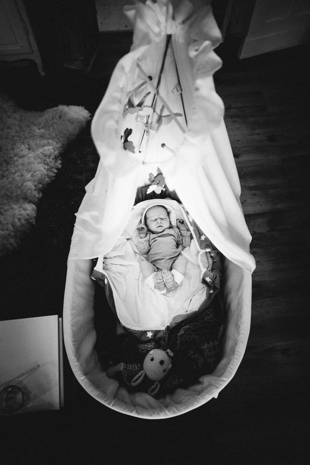 Eine Schwarz-Weiß-Aufnahem eines schlafenden Baby in einem klassischen Babybett mit Spielzeug und einer Mobile darüber.