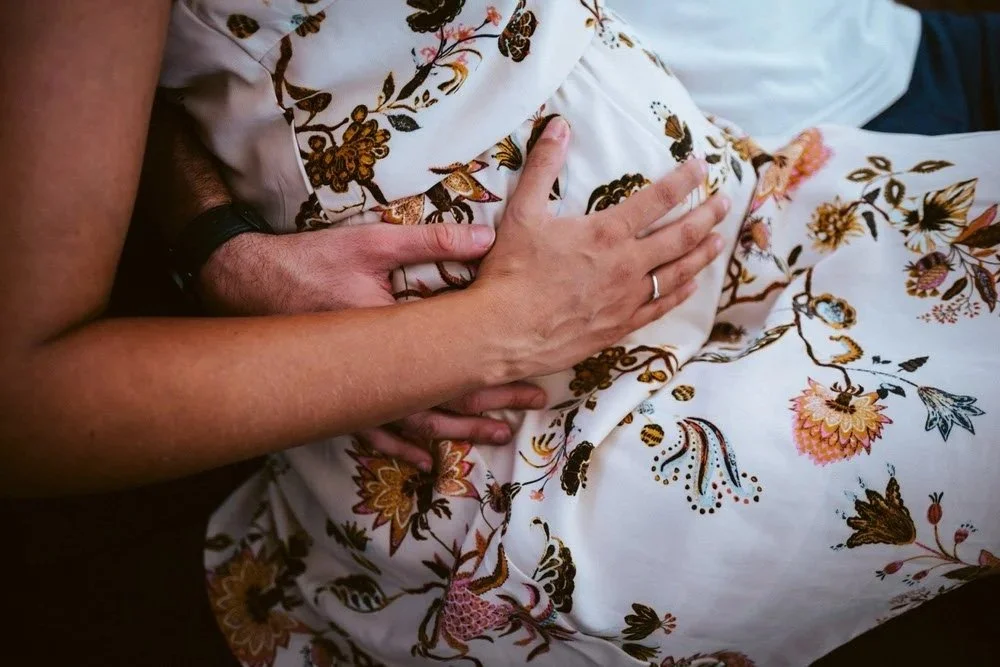 Nahaufnahme eines zärtlichen Moments von zwei Händen, die einander auf einem bunt gemusterten Kleid einer Frau mit Babybauch halten.
