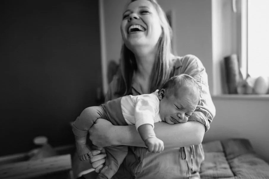 Eine lachende Mutter hält in dieser Momentaufnahme ein schlafendes Baby auf ihrem Arm. Die Szene ist in einem Raum mit einer Fensterbank mit Dekorationen. Das Bild ist schwarz-weiß.