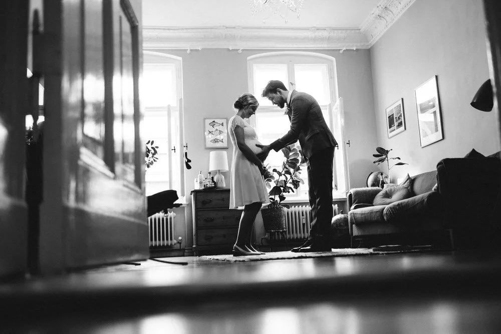 Ein Mann und eine Frau stehen in einem Wohnzimmer in Berlin, der Mann hält die Hände des Frau, das lächelt. Das Zimmer ist hell erleuchtet mit großen Fenstern, es gibt Pflanzen, Bilder an den Wänden und ein Sofa.