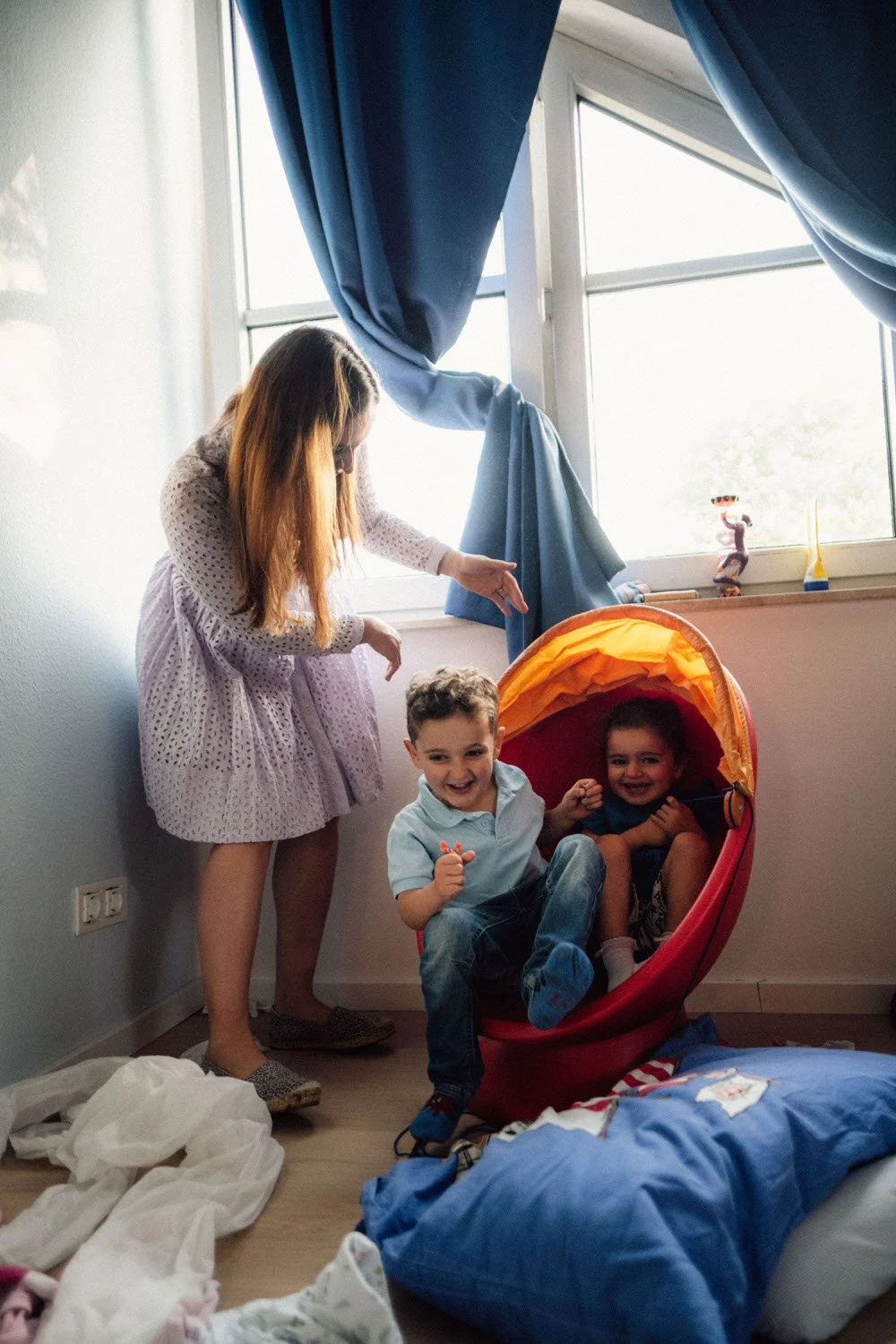 Eine Frau mit zwei Kindern, eines im Kinderbett und das andere im Sessel, spielen in einem Zimmer, das vollgepackt ist mit Wäsche und anderen Spielzeug. Es kommt stimmungsvolles licht durch die Fenster.