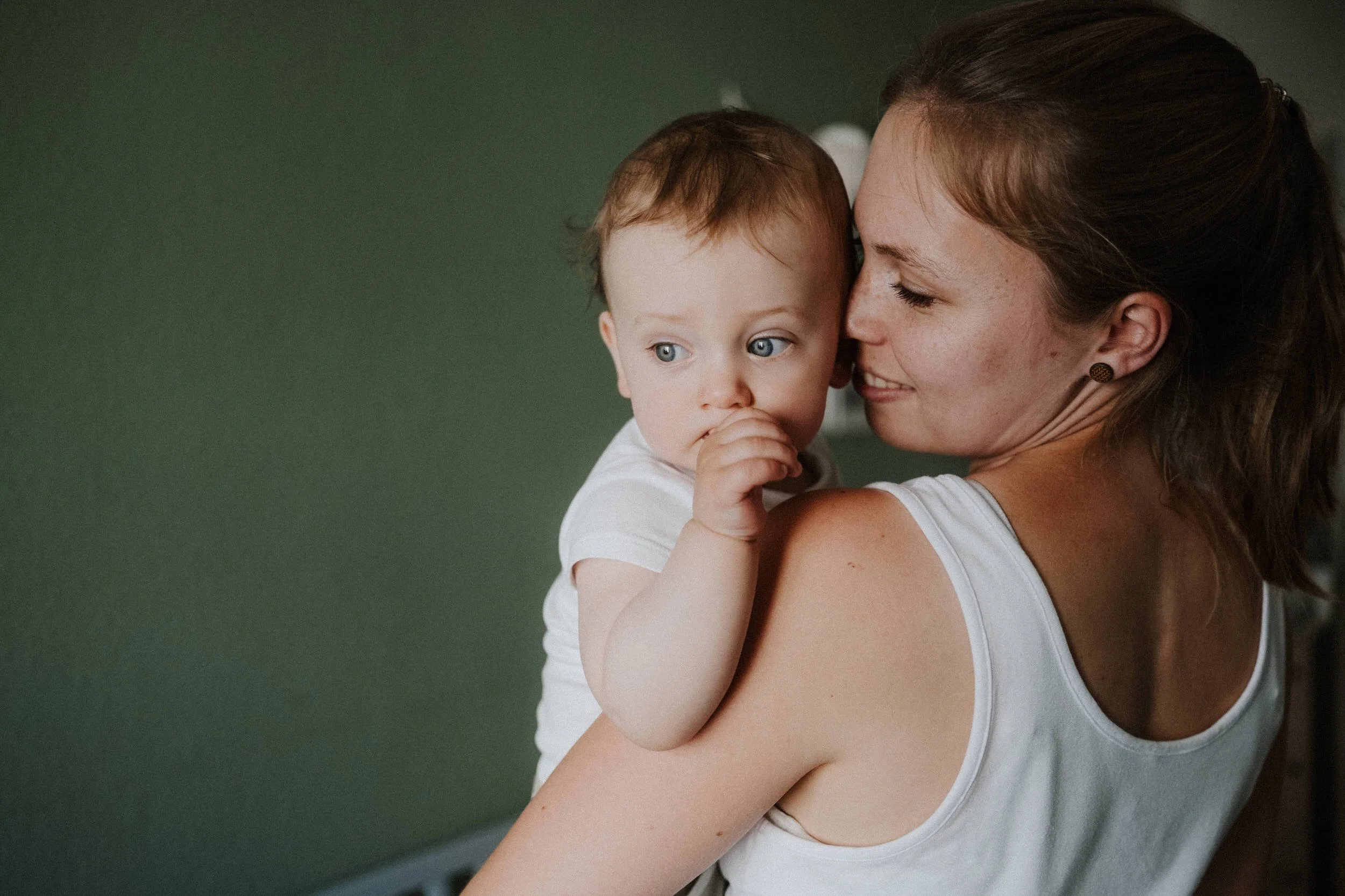Ein Moment zwischen Mutter und Sohn ganz persönlich und nah: Sie hält ihr Kind auf dem Arm und berührt es zärtlich mit der Nase. Beide tragen helle Kleidung und stehen bei Fensterlicht vor einer grünen Wand.