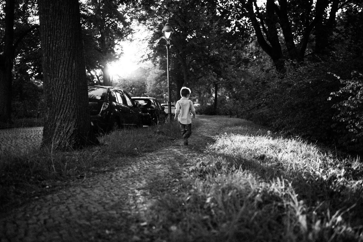 Ein junger Junge läuft auf einem schmalen Pfad durch einen Park bei Sonnenuntergang, umgeben von Bäumen und geparkten Autos, alles in Schwarz-Weiß.