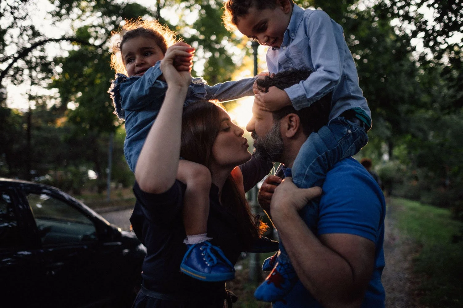 Familie, bestehend aus einem Mann, einer Frau und zwei Kindern, verbringt Zeit im Freien im Sonnenuntergang. Die Familie küsst sich liebevoll, während die Kinder auf den Schultern der Erwachsenen sitzen.
