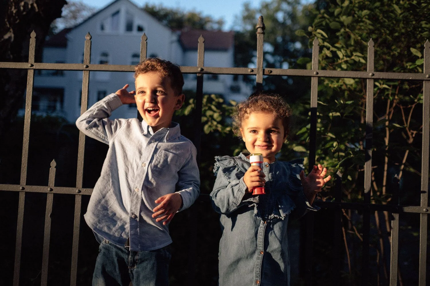 Zwei lachende Kinder stehen vor einem Gitterzaun, eines winkt und das andere hält Seifenblasen bei Dämmerlicht.