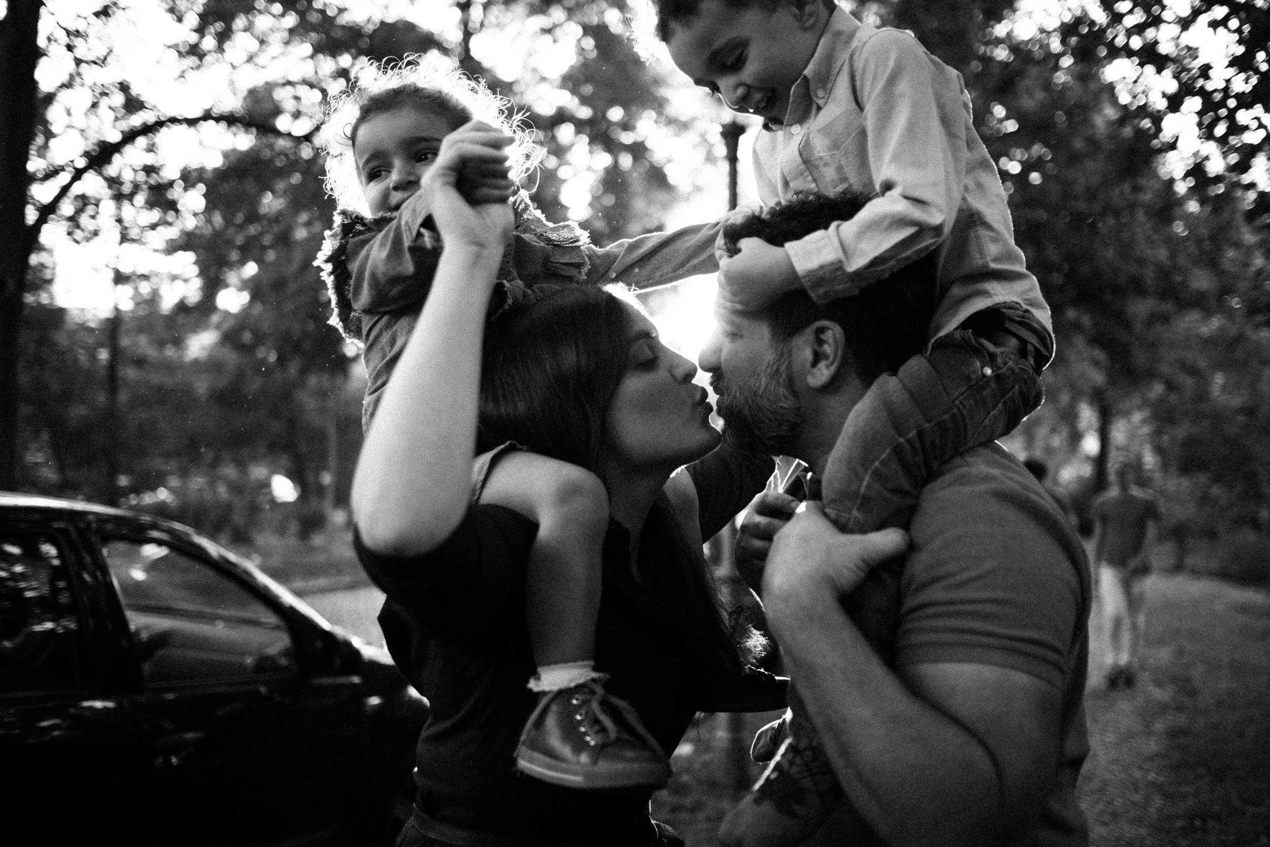 Ein Familienfoto in schwarz-weiß, bei dem ein Mann und eine Frau sich küssen, während zwei Kinder auf den Schultern der Erwachsenen sitzen und die Szene fröhlich wirkt.