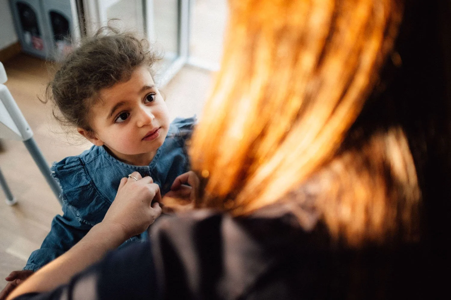 Ein kleines Mädchen mit lockigem Haar sieht aufmerksam auf eine seine Mutter mit roten Haaren, welche sie gerade anzieht.