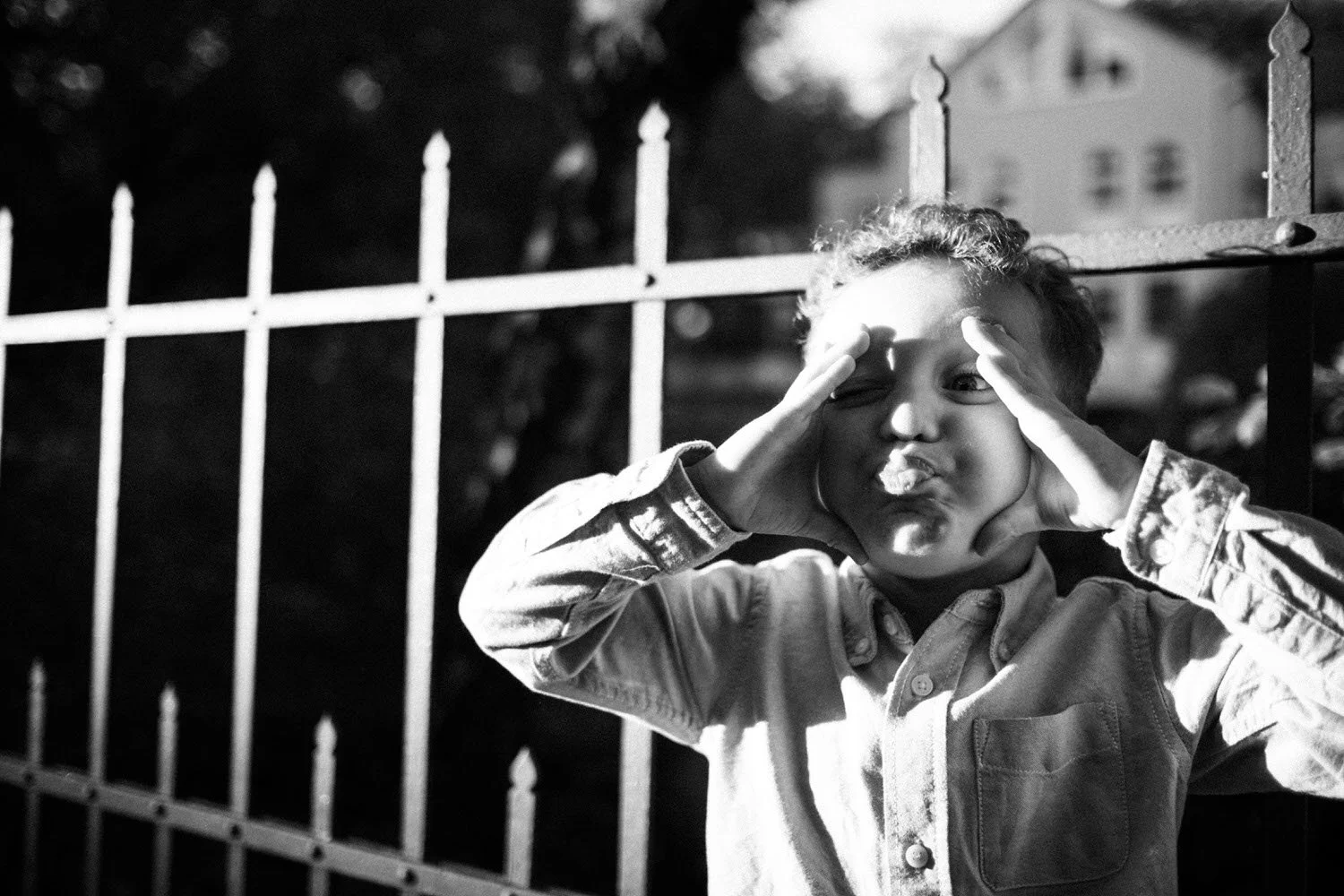 Junge macht eine Grimasse mit den Händen an am Gesicht und Zunge rausgestreckt vor einem Zaun im Freien in Schwarz-Weiß