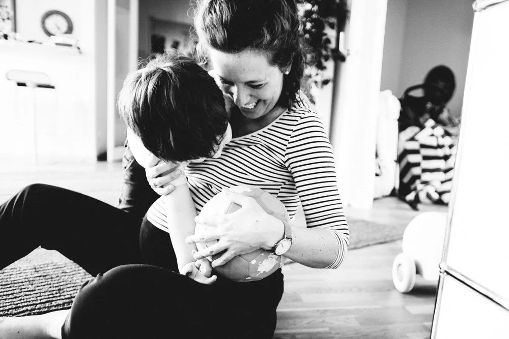 Eine schwangere Mutter in einer hellen Wohnung spielend und voller Freude mit ihrem Kind. Während dieser natürlichen Momentaufnahme hat sie ein Ball in der Hand, welchen sie von dem Kind zu verstecken versucht.