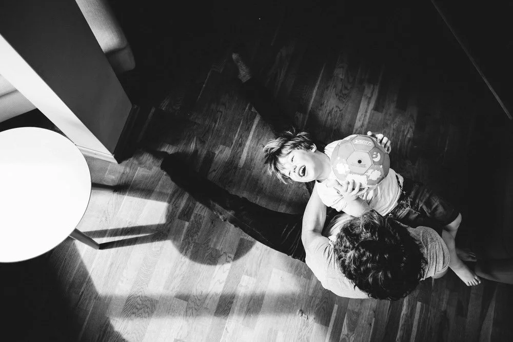 Ein Junge liegt in den Armen seines Vaters, hält einen Fußball und lacht laut, während beide spielen. Das Licht kommt stimmungsvoll in den Raum und leuchtet dem Kind ins Gesicht.