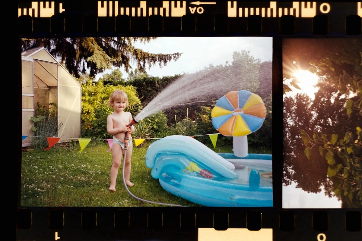 Ein Kind in Unterwäsche in einer analogen Fotografie, das im Garten mit einem Gartenschlauch Wasser sprüht, daneben ein aufblasbares Wasserbecken mit einem Sonnenschirm, bunten Flaggen und Bäumen im Hintergrund, bei Sonnenuntergang.