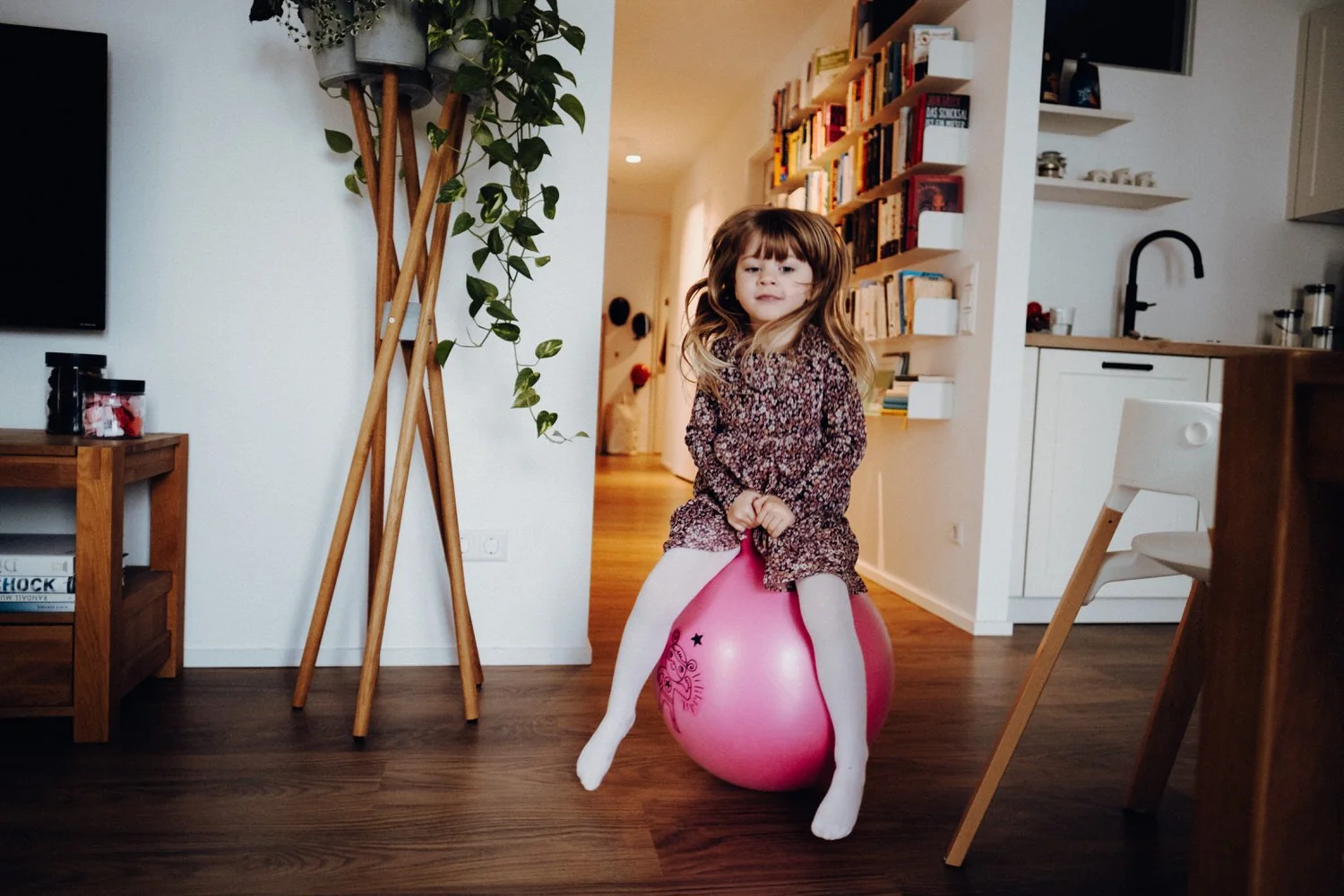 In der Alltagsszene sitzt ein Mädchen auf einem pinken Kinder-Hüpfball und springt damit durch die moderne Wohnung.