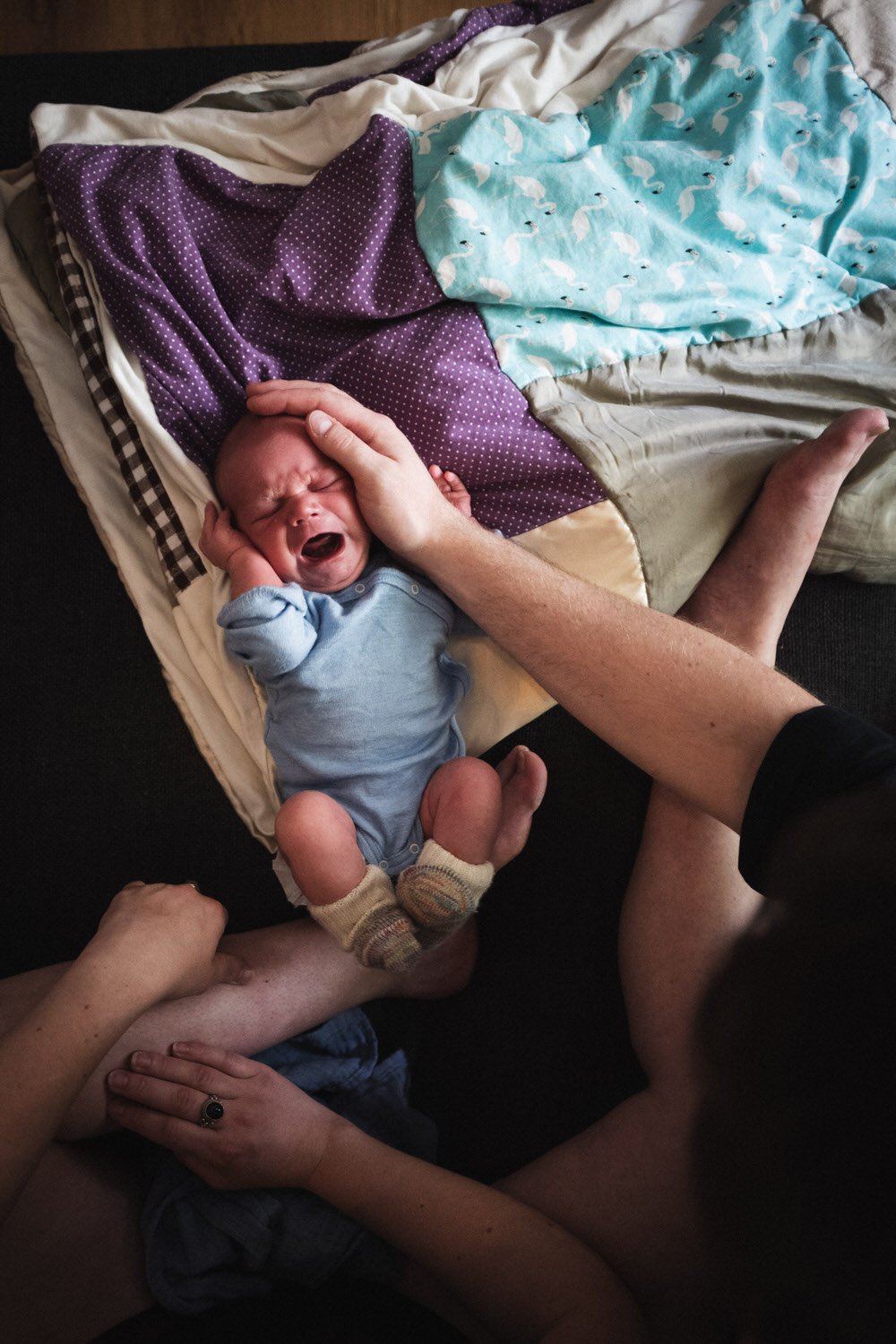 Authentischer Neugeborenen-Moment: Ein weinendes Baby, das von mehreren Erwachsenen gehalten wird, liegt auf einer Liege, umgeben von Decken.