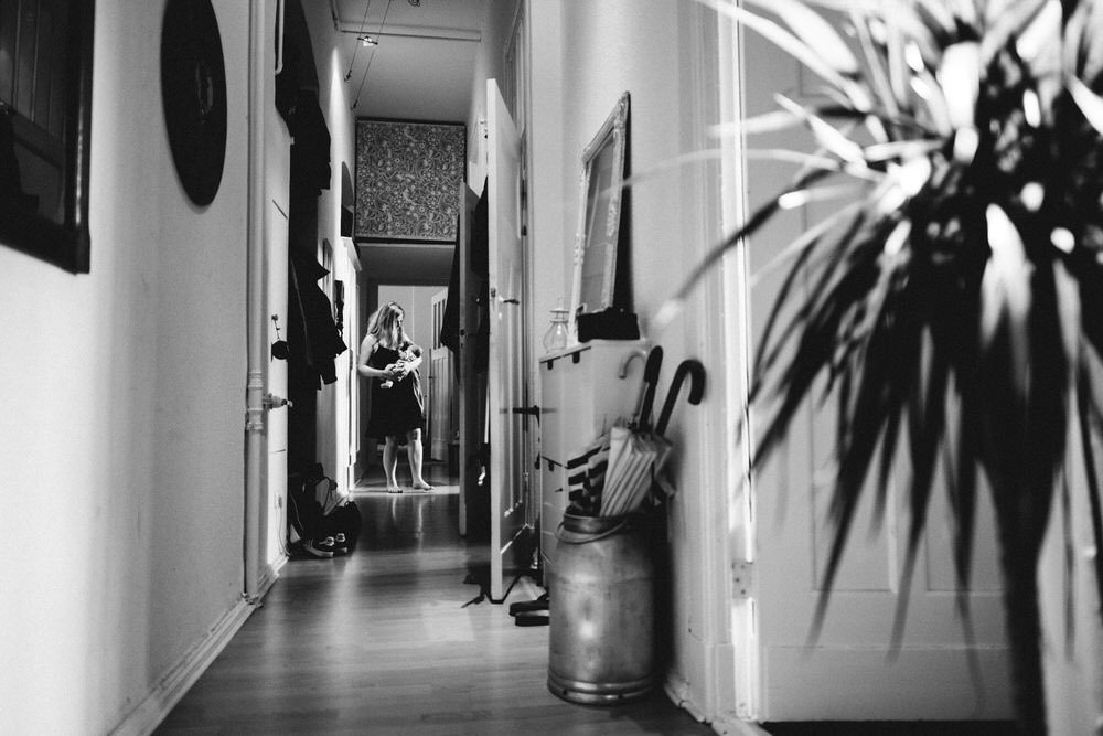 In einem langen Flur einer Altbauwohnung in Berlin mit einer Frau, die in der Ferne ein Baby hält. Die Aufnahme fängt die Umgebung detailliert mit ein. Im Vordergrund ist eine Zimmerpflanze zu erkennen.
