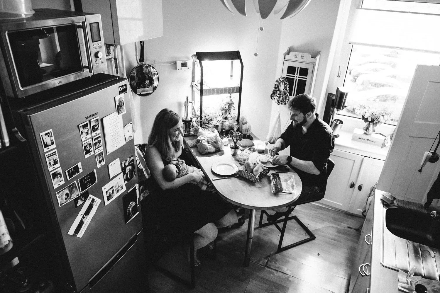 Authentischer Familienalltag: Eine Familie im Küchenraum, die Mutter hält ein Baby im Arm, während der Vater am Tisch das Essen zubereitet.