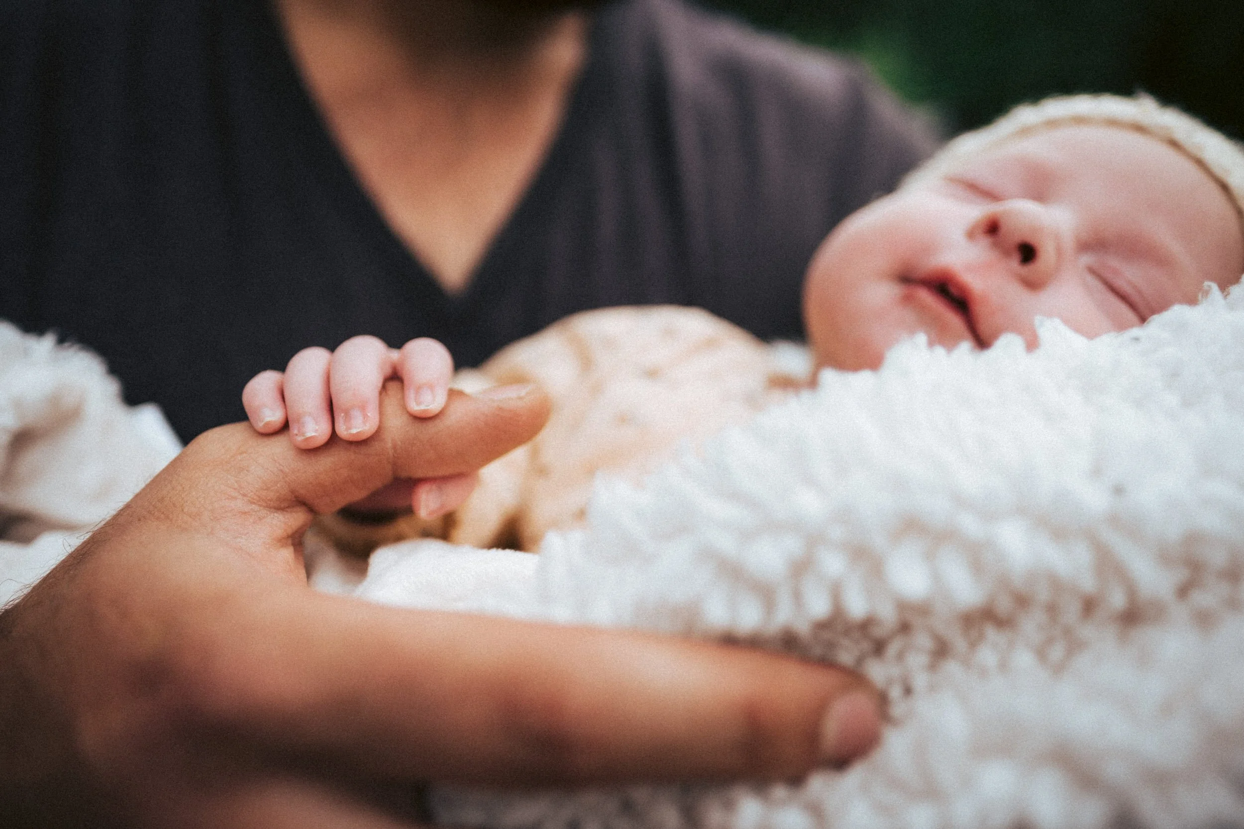 Die Fotoaufnahme zeigt eine große Männerhand an dessen Daumen von einem Baby bei einem Neugeborenenfotoshooting sanft festgehalten wird. 