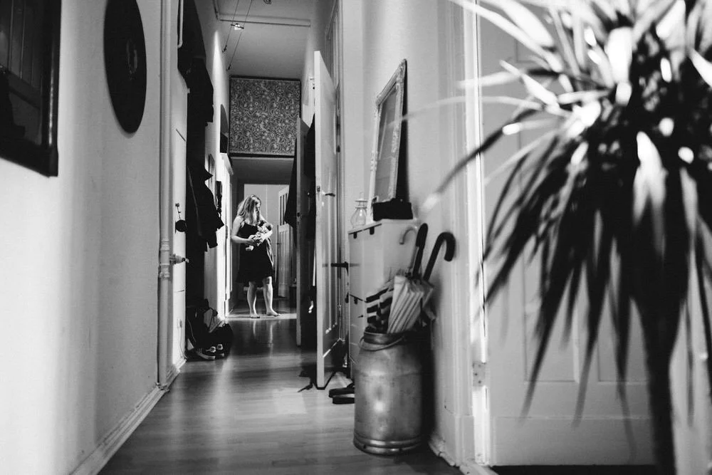 In einem langen Flur einer Altbauwohnung in Berlin mit einer Frau, die in der Ferne ein Baby hält. Die Aufnahme fängt die Umgebung detailliert mit ein. Im Vordergrund ist eine Zimmerpflanze zu erkennen.