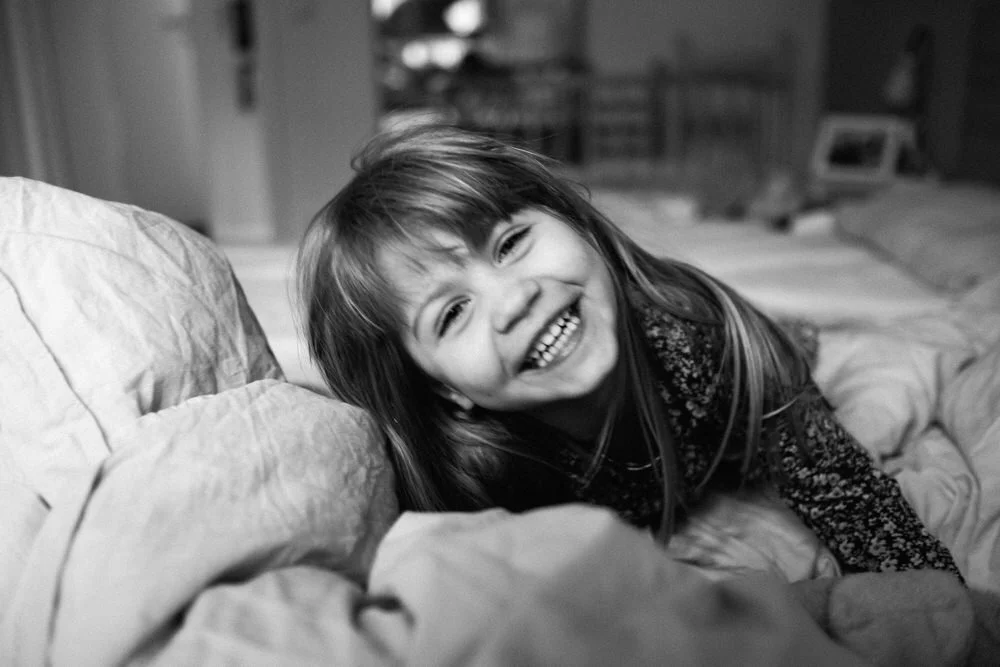 Ein Mädchen lachend und spielend auf einem Bett. Die schwarz-weiß Fotografie zeigt einen authentischen und kurzlebigen Moment.