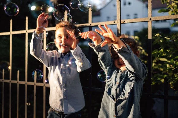 Zwei Kinder stehen vor einem Zaun mit warmen Sonnenlicht und fangen Seifenblasen. Es ist eine authentische Aufnahme voller Bewegung und Emotion