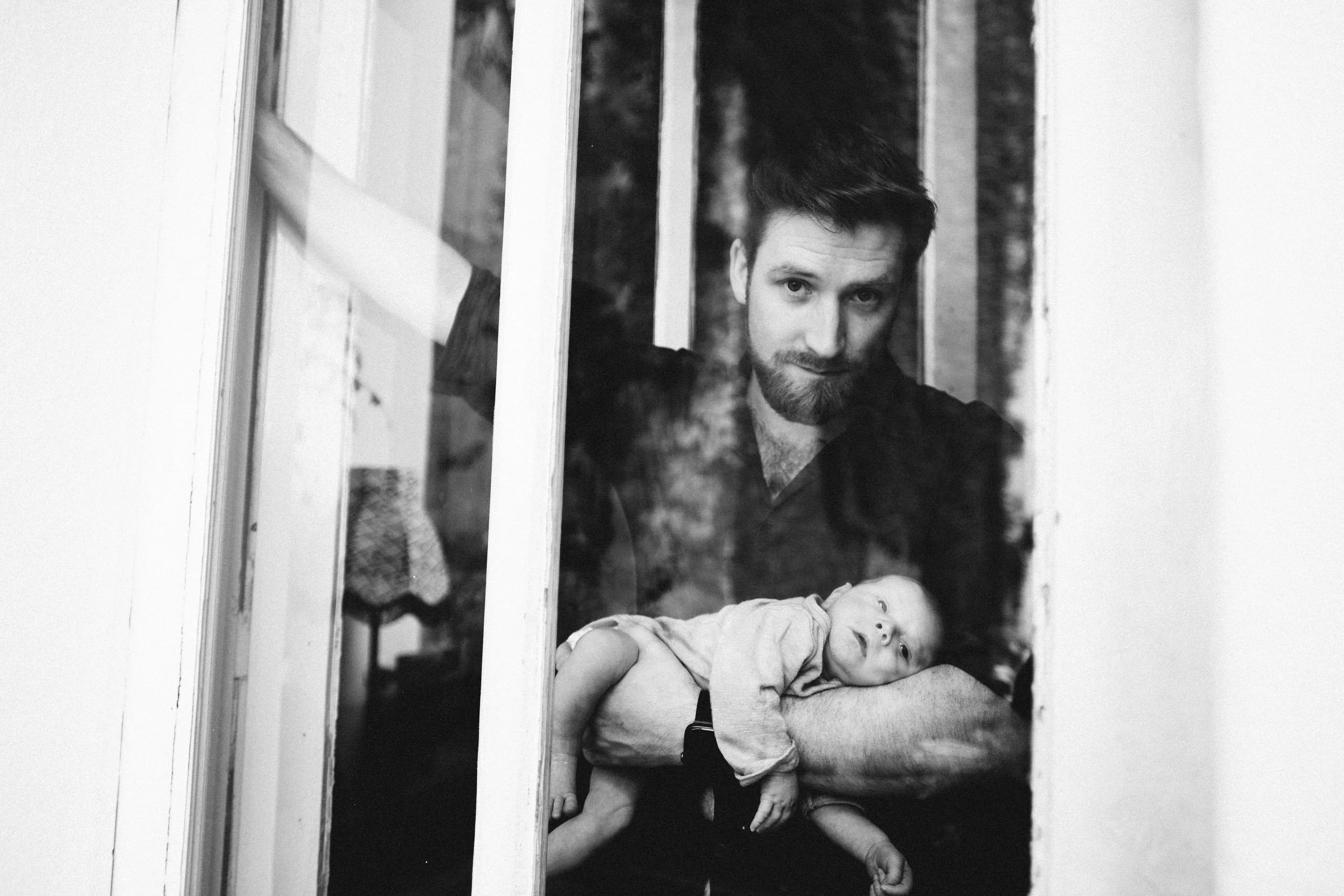 Ein junger Vater mit seinem Baby auf dem Arm schauen entspannt durch ein Balkonfenster. Bei dem Neugeborenenfotoshooting trägt der Vater trägt das Baby auf einem Arm und schaut stolz in die Kamera.