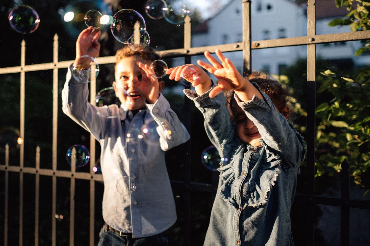 Zwei Geschwister in einer authentischen Momentaufnahme, spielen mit Seifenblasen vor einem Zaun bei Sonnenuntergang, lachen und haben Spaß.