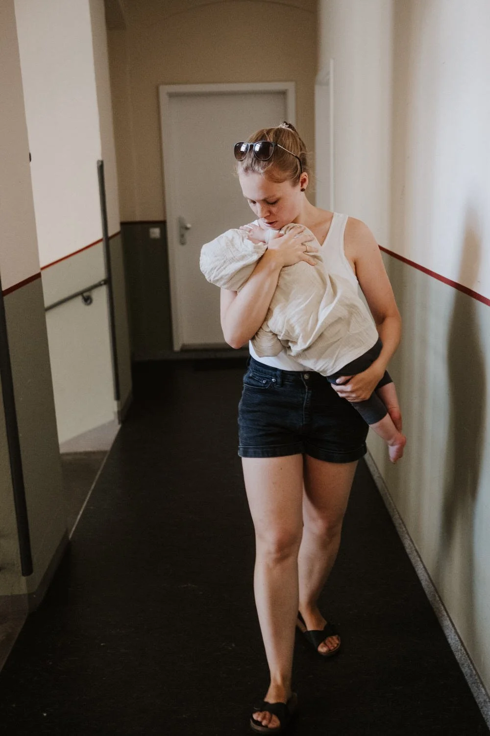Eine junge Frau trägt ein schlafendes Baby in den Armen, während sie durch einen Flur in einem Berliner Altbau geht.