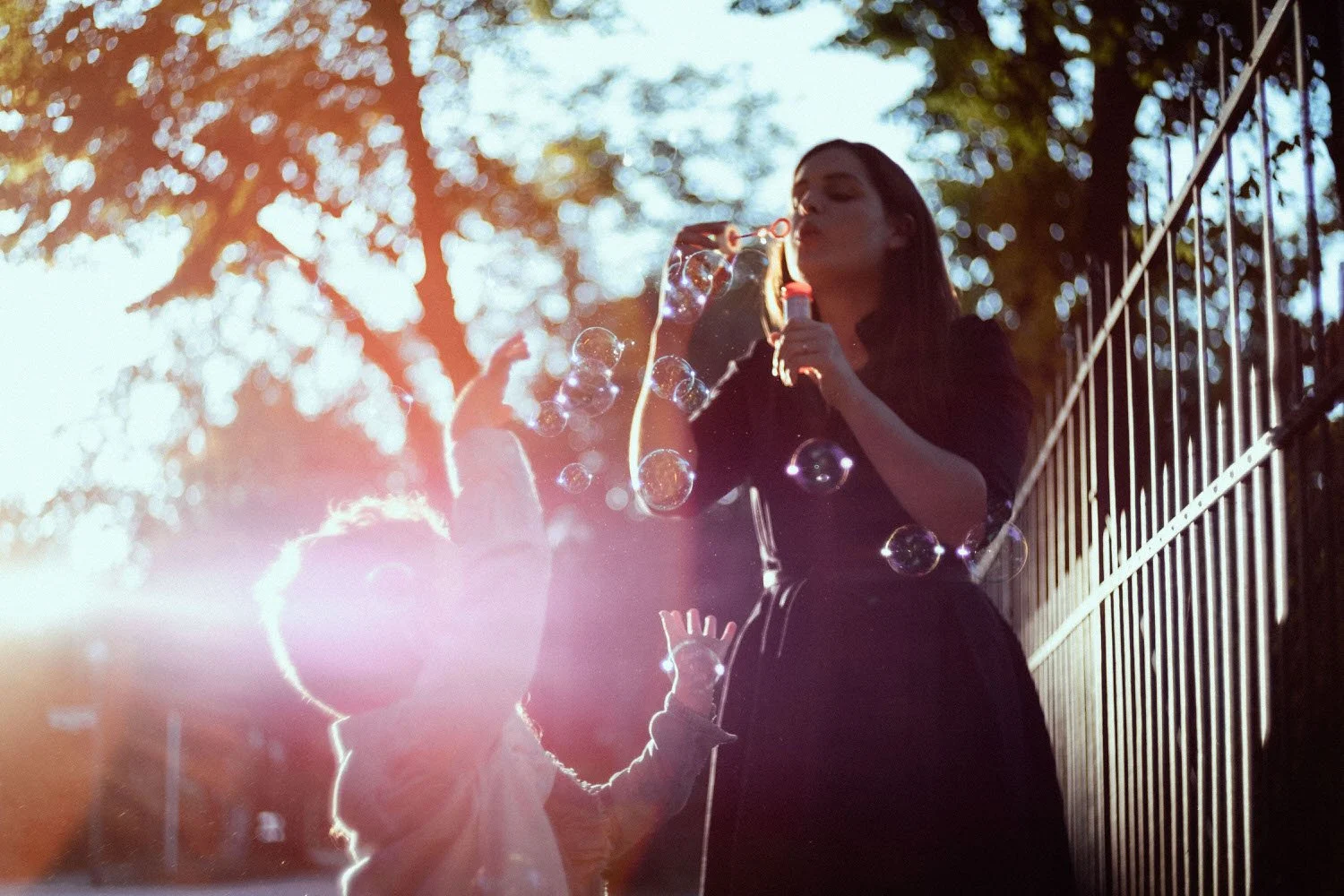 Familiensituation in der Freizeit: Eine junge Frau bläst Seifenblasen im Freien bei Sonnenuntergang, während ein Kind sie beobachtet und die Seifenblasen fangen. 