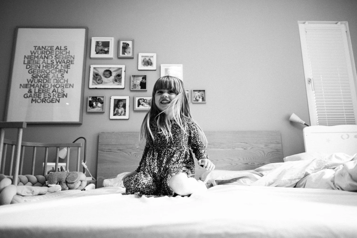Kleines Mädchen mit langem Haar in einer Momentaufnahme lachend auf einem Bett in einem Schlafzimmer, schwarz-weiße Aufnahme.