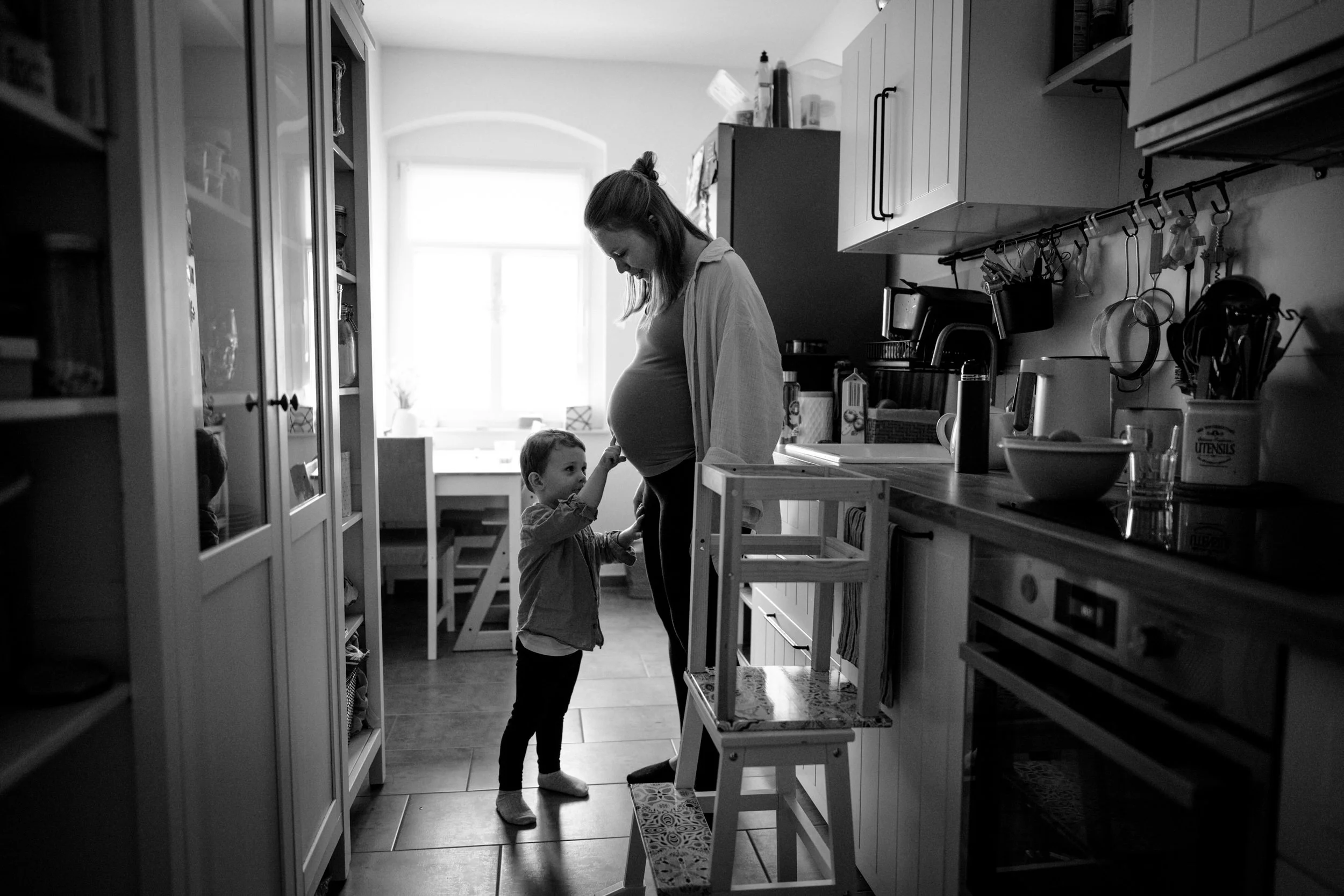 Frau mit Babybauch und Sohn in der Küche, die sich anlächelnd zum Bauch der Frau vorsichtig berühren. Es handelt sich um eine Schwarz-Weiß-Aufnahme