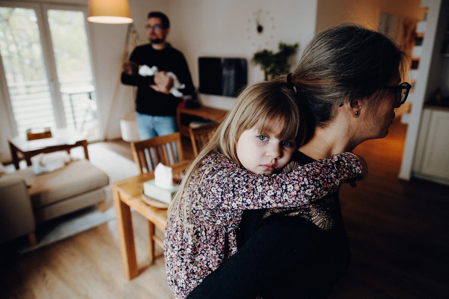 Ein Mädchen mit blonden Haaren und blauen Augen liegt auf der Schulter einer Frau, das Mädchen schaut verträumt. Im Hintergrund steht ein Mann, der ein kleines Baby hält. Sie stehen im Wohnbereich einer modernen Berliner Wohnung.