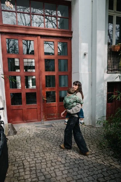 Eine Frau trägt ein Kind, während sie vor einer roten Holztür im Außenbereich eines Hauses steht. Es ist ein sonniger Tag, und die Frau lächelt. Die Umgebung ist mit Pflanzen und Steinen gepflastert.