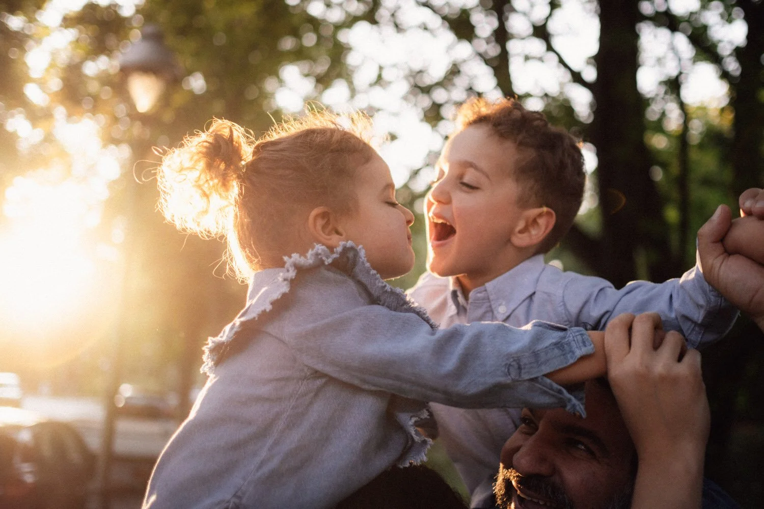 Zwei Kinder, ein Junge und ein Mädchen, spielen lachend im Freien bei Sonnenuntergang, während sie auf den Schultern der Eltern getragen werden.