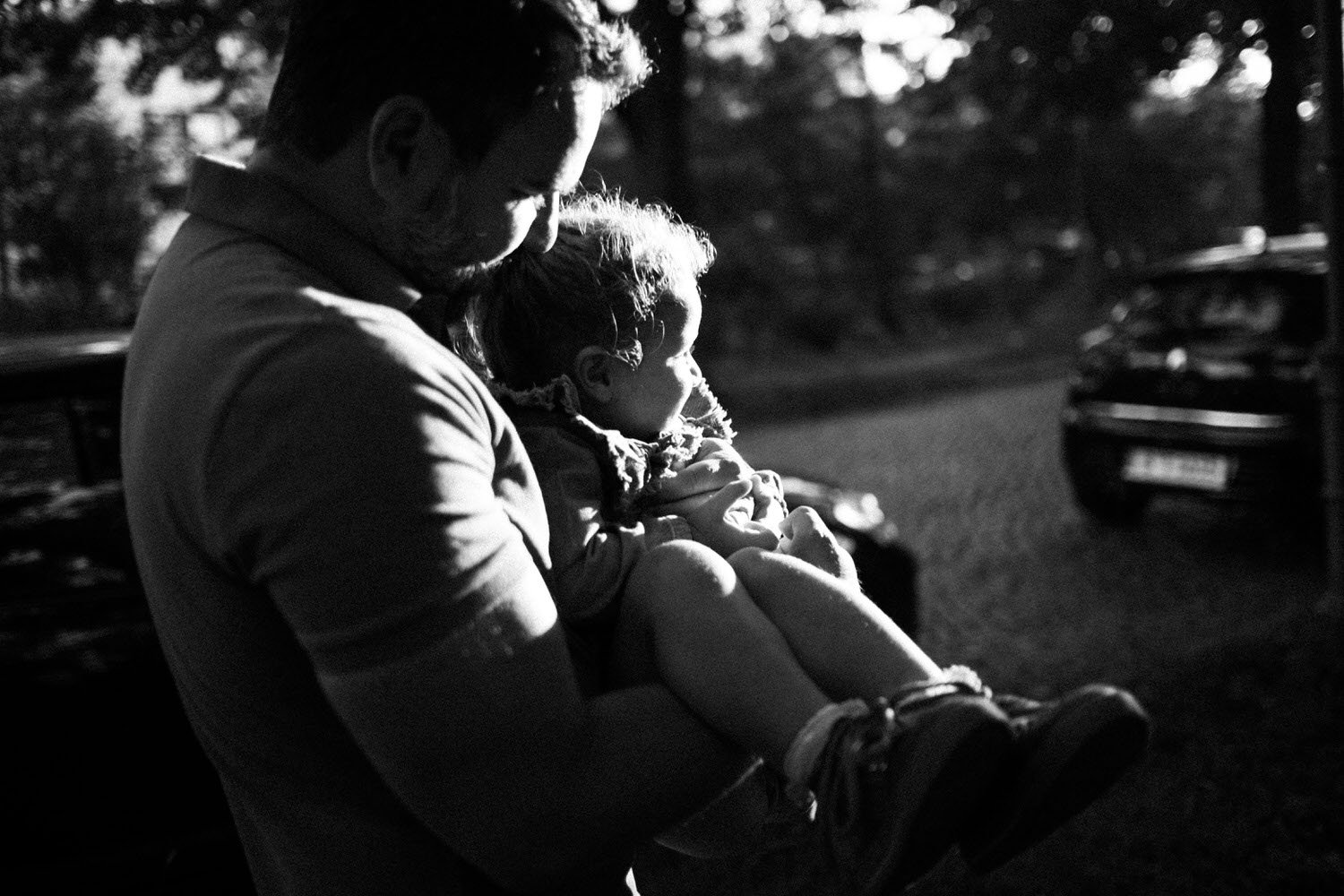Ein Mann hält ein Kind auf dem Schoß, beide schauen in die Sonne. Es ist eine schwarze und weiße Aufnahme, die im Freien in einer West-Berliner Alle aufgenommen wurde.