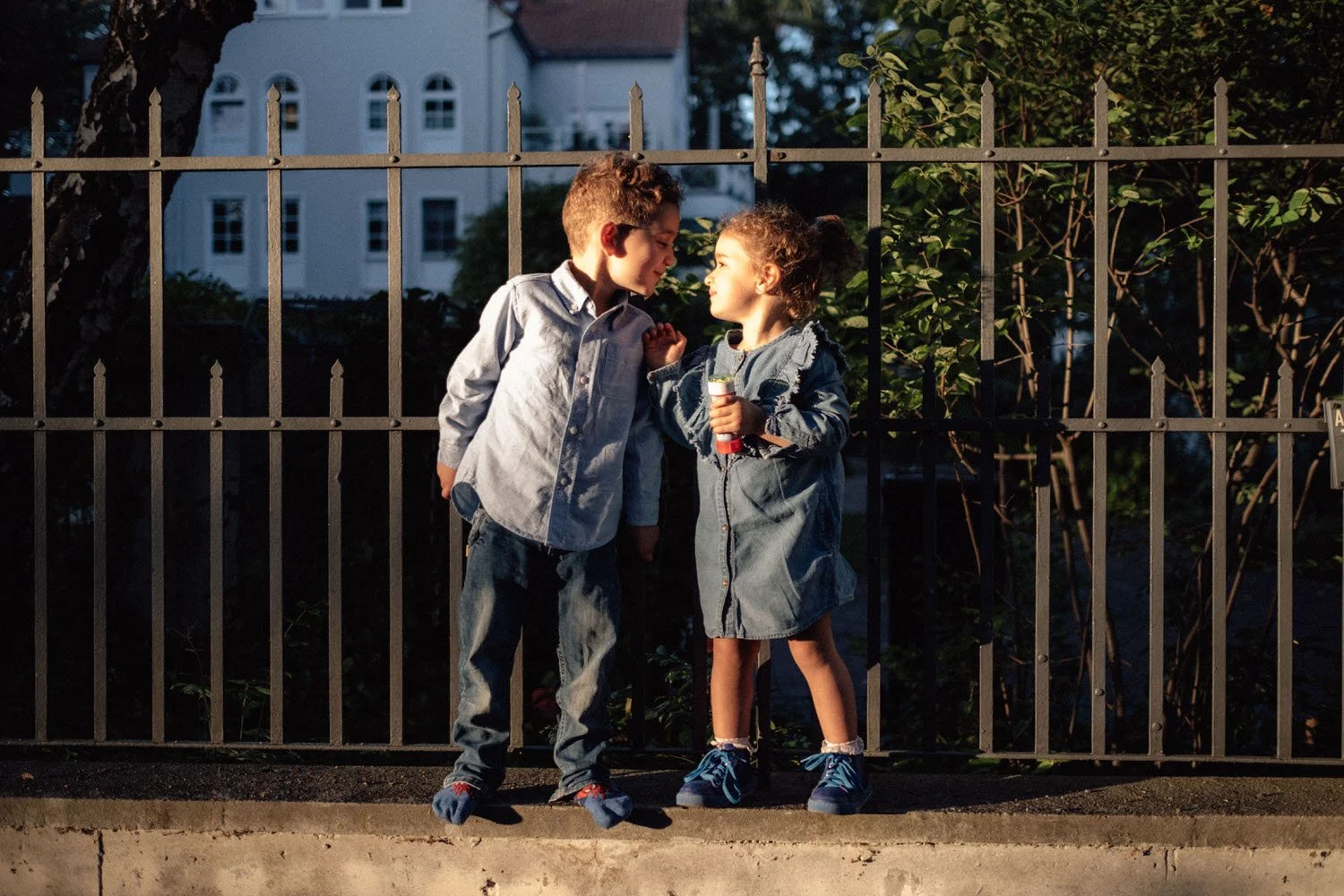 Zwei Kinder, ein Junge und ein Mädchen, stehen dicht beieinander und schauen in die Augen an einem Metallzaun im Abendlicht.