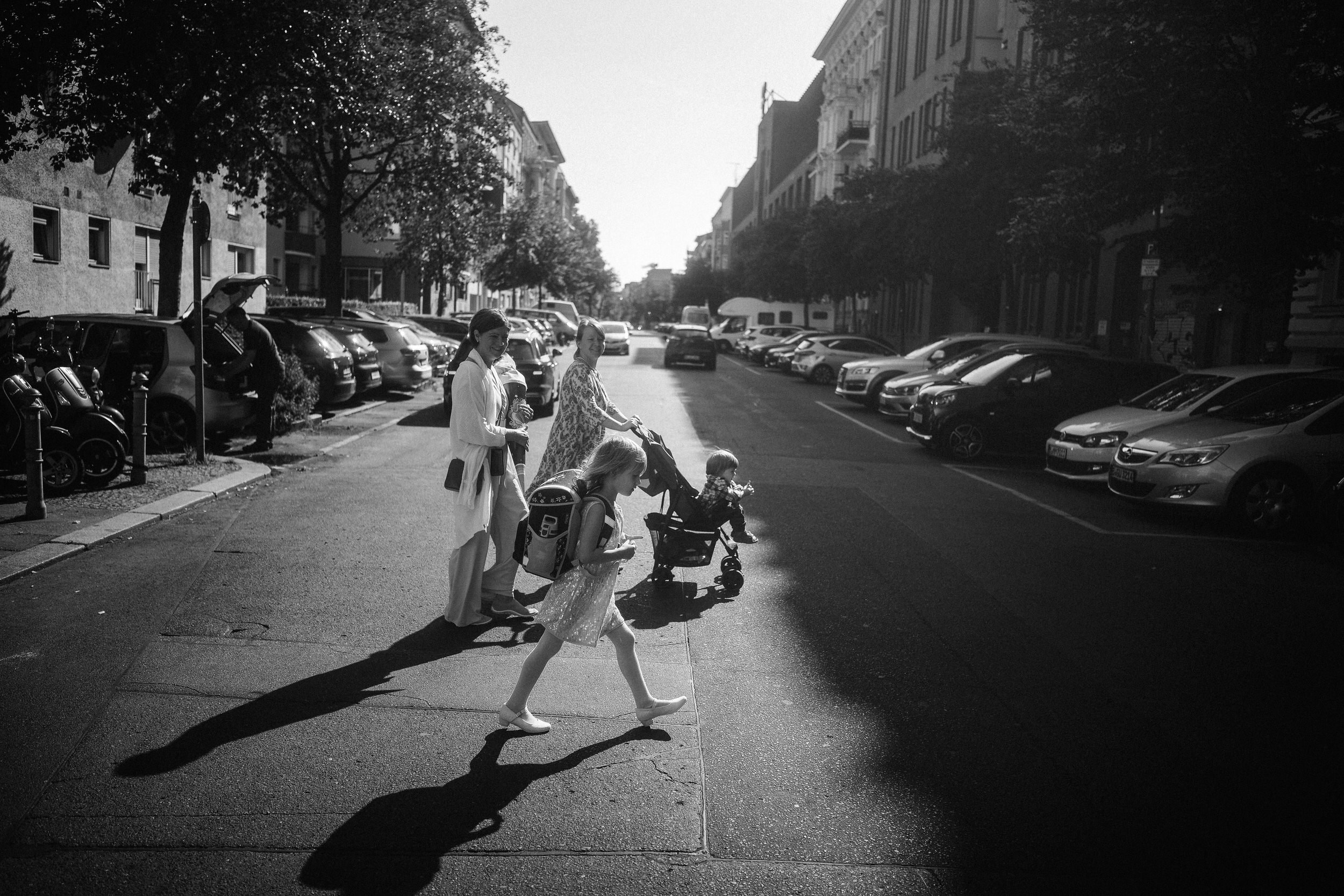 In eine Szene mit Gegenlicht laufen ein Kind mit Schultasche und eine kleine Gruppe von Menschen über eine Straße. Diese Familienreportage entstand während einer Einschulung. 