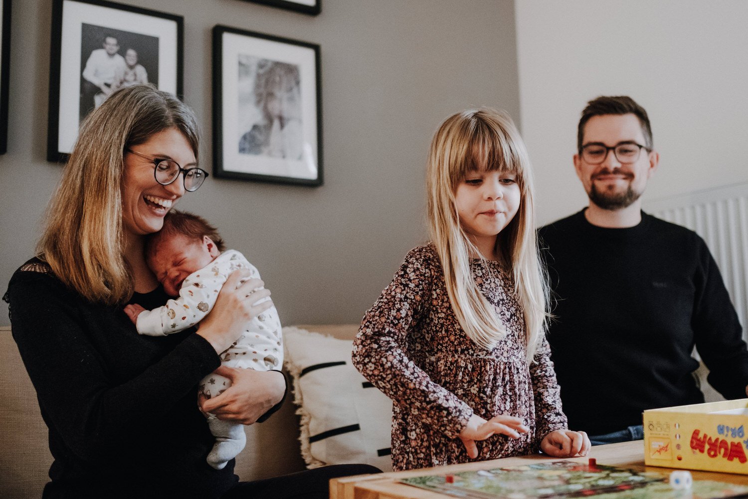 Familie bei einem Brettspiel-Nachmittag, Mutter hält Baby, Tochter spielt, Vater schaut und lächelt.