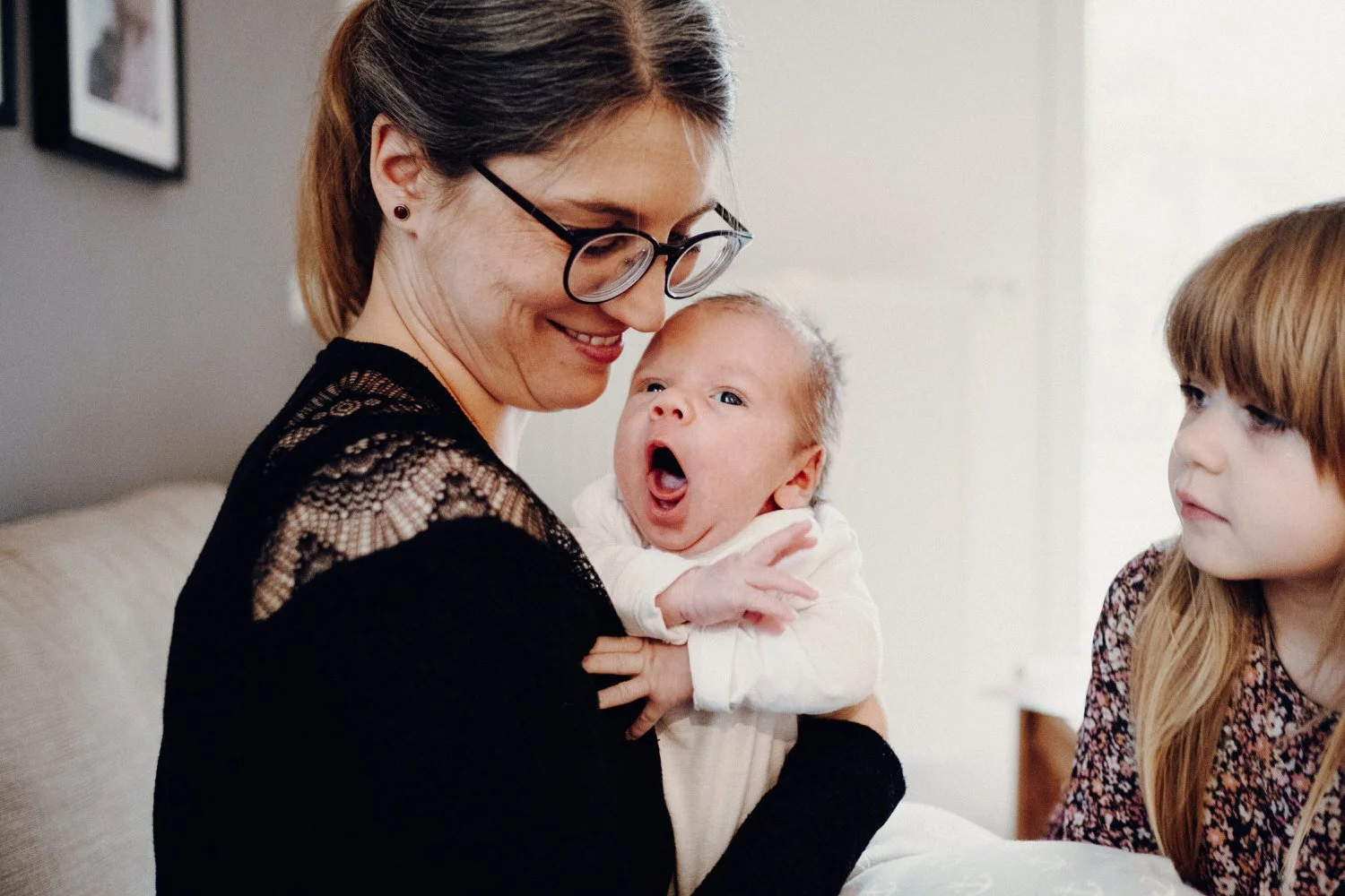 Eine Mama hält ein gähnendes Baby im Arm, während seine Schwester zusieht.